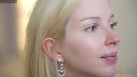 Video of a fair-skinned, blonde woman with blue eyes and subtle makeup, wearing a heart-shaped earring. Background is blurred, focusing on her profile.