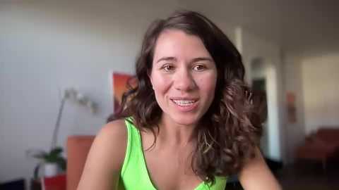 Media: Video of a smiling, middle-aged woman with curly brown hair, wearing a bright green tank top, indoors, with a blurred background featuring a white wall and a plant.