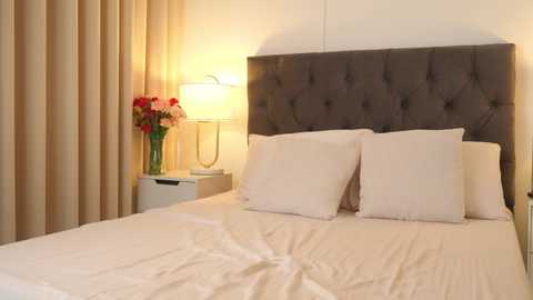 Media: Video of a modern bedroom with a tufted gray headboard, white pillows, and beige curtains. A white nightstand holds a vase with red and pink flowers, a lamp, and a black remote control.