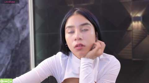 Media: Video of a young Asian woman with straight black hair, wearing a white long-sleeve top, sitting with her head resting on her hand, looking tired, in a modern room with dark walls and a staircase.