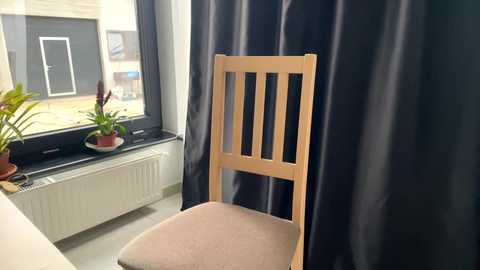 Media: Video of a minimalist room with a wooden chair and dark curtains. Potted plants and a radiator are visible on a window ledge, and light streams through the window, highlighting the room's simple, modern design.