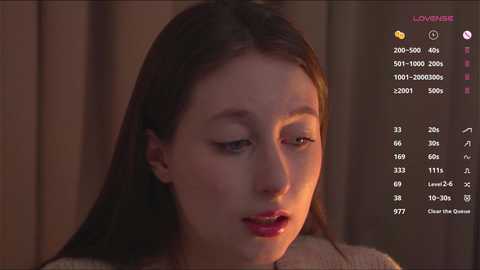 Video of a young Asian woman with straight brown hair and light skin, wearing a beige sweater, looking pensive in a dimly-lit room. A social media app interface with her stats is visible.