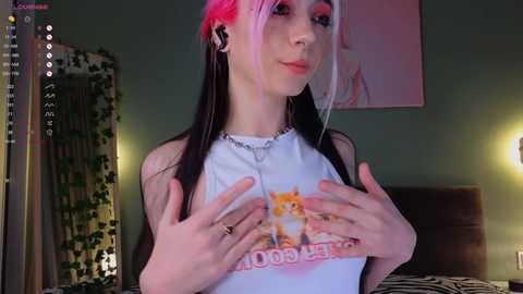 Media: Video of a young woman with fair skin and a white tank top with a colorful graphic, wearing black and pink hair extensions and silver hoop earrings, in a dimly lit bedroom.