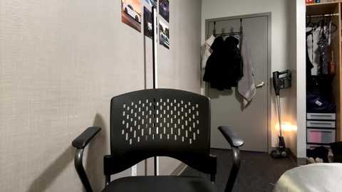 Media: Video of a black mesh-back chair with armrests in a small, cluttered room with beige walls, a door, and a clothes rack with hanging items, a lamp, and a floor lamp.