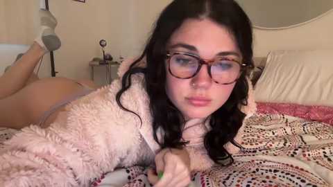 Media: Video of a young woman with fair skin, long dark hair, and glasses lying on a bed, wearing a fluffy, light pink sweater and beige underwear, surrounded by patterned bedding.