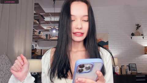 Video of a young Asian woman with long black hair, wearing a white cardigan, holding a blue iPhone, smiling in a cozy, modern living room with white brick walls, bookshelves, and a lamp.