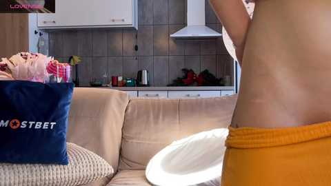 Video of a nude man in orange shorts, standing in a modern kitchen with beige couch, blue bag, and white table.