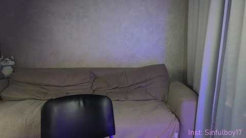 Media: Video of a dimly lit, cluttered room with a beige sofa and a black chair, featuring a grayish wall and sheer white curtains.