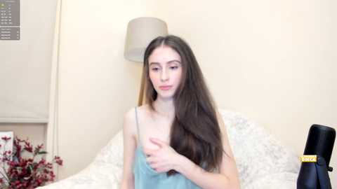 Media: Video of a young, slim Caucasian woman with long, straight brown hair, wearing a light blue camisole, seated on a bed with white sheets, in a beige room with a lamp and a vase of red flowers.