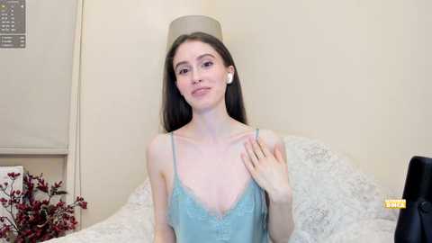 Media: Video of a slender, fair-skinned woman with long dark hair, wearing a light blue lace camisole, white earbuds, and a beige lampshade hat, seated on a white bed with floral pillows.