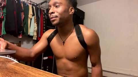 Media: Video of a shirtless, muscular Black man with short hair, wearing a black harness and necklace, standing in a cluttered closet with hanging clothes.