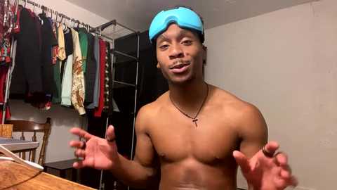 Media: Video of a shirtless, muscular Black man with a blue headband, wearing a necklace. He's in a cluttered room with hanging clothes and a wooden chair.