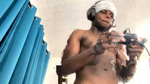 Media: A topless, muscular Black man with a beard and white hat, wearing headphones, holds a camera in a dimly-lit room with blue curtains.