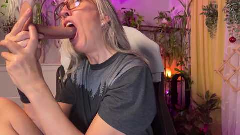 Media: Video of a blonde woman with glasses, wearing a black t-shirt, performing oral sex on a large, erect penis. Background features green plants and a yellow curtain.