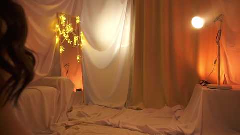 Media: Video of a dimly lit, romantic setup with white drapes, a glowing yellow lantern, and warm orange light from a table lamp. The scene exudes a soft, intimate ambiance.