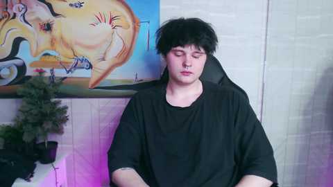 Media: Video of a young man with dark hair, wearing a black t-shirt, sitting in a black gaming chair. Background features a colorful abstract painting and a potted plant.