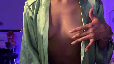 Media: Video of a woman with light skin, wearing an unbuttoned green satin robe, revealing her medium-sized breasts. Her right hand is covering her left breast. Background shows a dimly lit room with purple lighting.