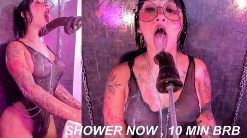 Media: Video of a tattooed woman with short black hair, wearing glasses and a black bodysuit, licking a black dildo attached to a chain in a shower. Text reads \"SHOWER NOW 10 MIN BRR.\