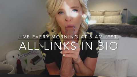 Video of a blonde woman with blue eyes performing oral sex on a large, erect penis in a bedroom setting. Text overlays read \"Live Every Morning at 5 AM (EST) ALL LINKS IN BIO.\