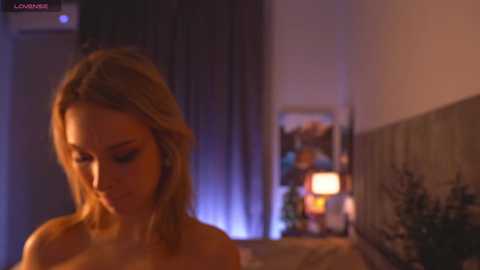 Media: Video of a blonde woman with medium-length hair, topless, in a dimly lit bedroom with a warm, cozy ambiance.