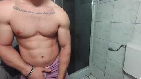 Media: Video of a shirtless man with a muscular chest, a tattoo above his nipples, and a purple towel around his waist, standing in a tiled bathroom with a shower and a toilet roll holder.