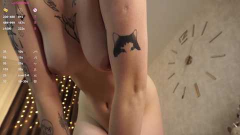 Media: Video of a light-skinned, nude woman with medium-sized breasts, inked with a black cat tattoo on her shoulder, and a large clock on the wall.