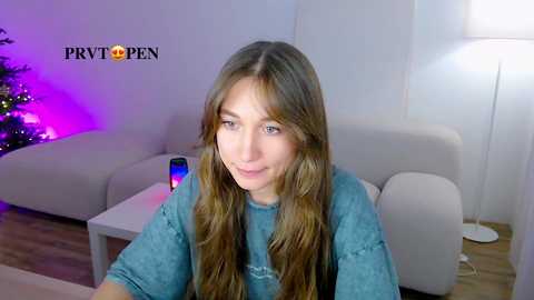 Media: Video of a young woman with long, wavy brown hair, wearing a blue t-shirt, sitting on a white couch in a modern living room with a Christmas tree, purple lights, and a lamp.