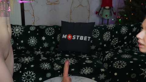 Media: A video of a woman with light skin, wearing a black top, sitting on a dark green couch with snowflake patterns. A man's erect penis is in the foreground. Background includes a Christmas tree and a black pillow with white snowflakes.