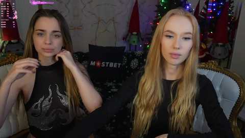 Media: Video of two young women with long hair, one brunette and one blonde, wearing black tops, seated on a couch with Christmas decorations in the background.