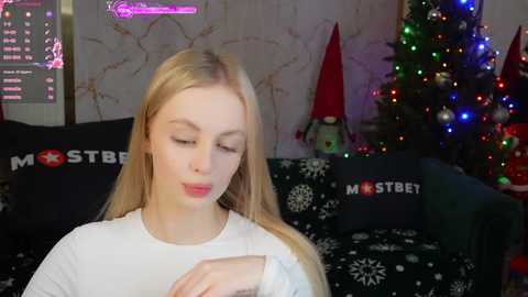 Media: Video of a young, fair-skinned woman with long blonde hair, wearing a white long-sleeve shirt, sitting on a black couch adorned with white snowflakes and \"MOSTBEET\" text. The background features a decorated Christmas tree with colorful lights, a red hat, and a plush toy.