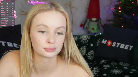 Media: Video of a young, fair-skinned, blonde woman with straight hair, wearing a top, sitting on a dark sofa with white snowflake pattern. Background features a Christmas tree with colorful lights and red decorations.