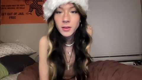 Media: Video of a young, fair-skinned woman with long, wavy hair, wearing a Santa hat, a nose ring, and a necklace, in a bedroom with beige walls and a bed.