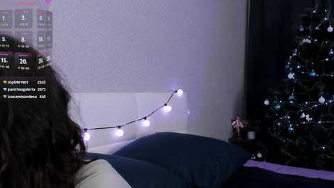 Video of a woman with long curly hair, lying on a bed, watching a calendar on a white wall with a Christmas tree adorned with blue lights and white ornaments.