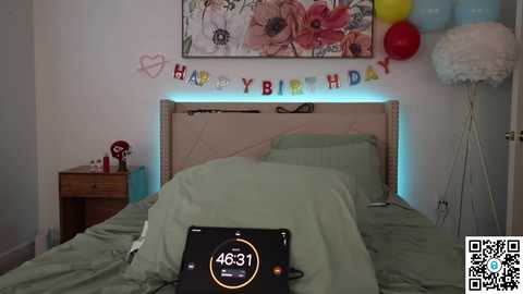 Media: Video of a bedroom with a white wall, a wooden headboard, green bedspread, and a digital clock reading 4631. A birthday banner hangs above, balloons and a stuffed animal are present, and a QR code is visible.