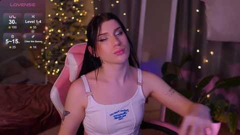 Media: Video of a young woman with fair skin and dark hair, wearing a white tank top, sitting in a pink gaming chair. Background features a cozy room with string lights, plants, and a TV.