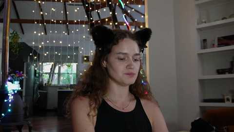 Media: Video of a young woman with curly hair wearing black cat ears and a black sleeveless top, indoors with fairy lights, bookshelves, and a cozy atmosphere.