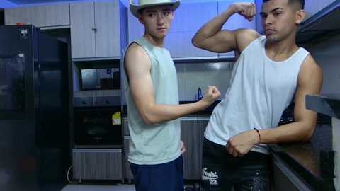 Media: Video of two young men, one flexing bicep, wearing tank tops, baseball caps, and jeans, in a modern kitchen with stainless steel appliances.