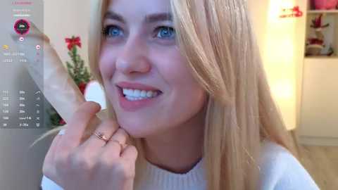 Media: A candid video of a smiling, fair-skinned blonde woman with blue eyes, wearing a white sweater, holding a large, realistic-looking dildo. The background features a Christmas tree and shelving with gifts.