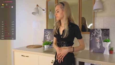 Media: Video of a blonde woman in a black crop top and high-waisted pants, standing in a modern kitchen with beige tiles, potted plants, and a black-and-white framed photo on the counter.