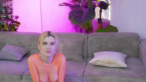 Media: Video of a fair-skinned, blonde woman with a slender physique, wearing a sheer, orange top, seated on a gray-patterned couch. Background features green plants and purple lighting.