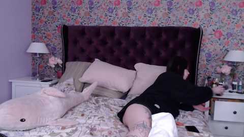 Media: Video of a woman in black lingerie lying on a floral-patterned bed with a plush shark, purple headboard, and pink pillows.