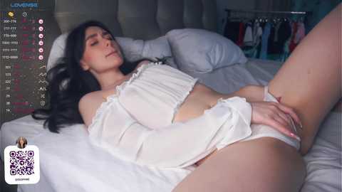 Media: Video of a slender, light-skinned woman with long black hair, wearing a sheer white crop top and panties, lying on a bed with white sheets.