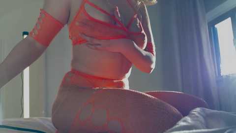 Media: Video of a fair-skinned woman in red lingerie, kneeling on a bed, covering her breasts with one hand, in a softly lit, modern bedroom with white curtains.