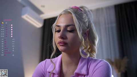 Media: A video of a young Caucasian woman with blonde hair tied back, wearing a light purple shirt, looking contemplative in a modern, dimly lit room with a calendar and air conditioning unit visible.