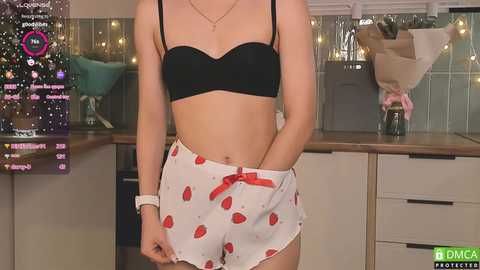 Media: Video of a slender woman in a black bra and white shorts with red strawberries, standing in a modern kitchen with tiled backsplash, a vase, and a counter.