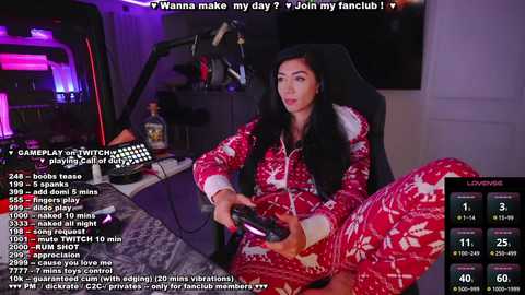 Media: Video of a woman in a red, reindeer-print onesie, sitting on a gaming chair, holding a gaming controller, in a dimly-lit room with a large TV and gaming setup.