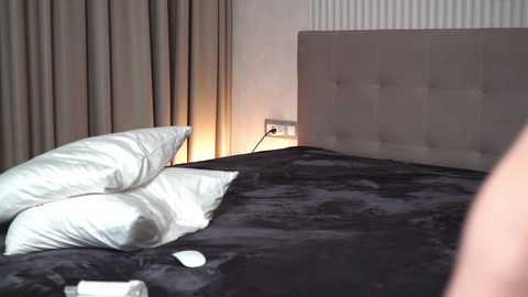 Media: Video of a modern bedroom with a dark, plush bedspread, white pillows, beige curtains, and a light gray upholstered headboard.