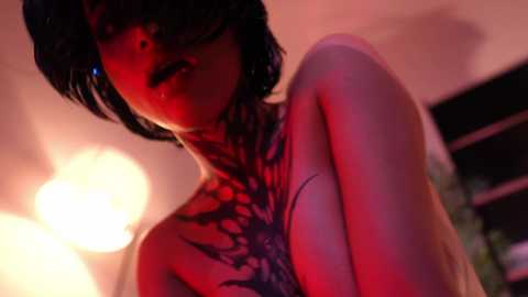 Media: A video of a topless woman with short black hair and a tattoo covering her chest, bathed in red and orange lighting.