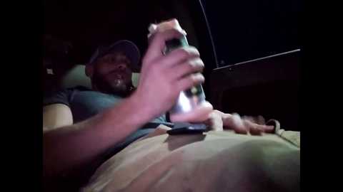 Media: Video of a man with a beard and a cap, holding a beer bottle, sitting in a dimly lit car, wearing a gray t-shirt and beige shorts.