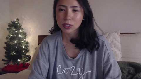 Media: Video of a young Asian woman with long black hair, wearing a cozy gray \"Cozy\" sweatshirt, seated indoors with a lit Christmas tree and festive decorations in the background.